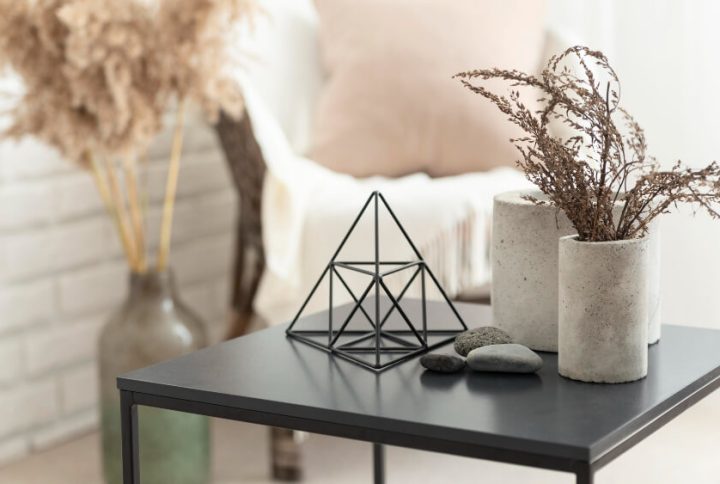 A minimalist black side table holding a geometric wire sculpture and concrete vases with dried plants..