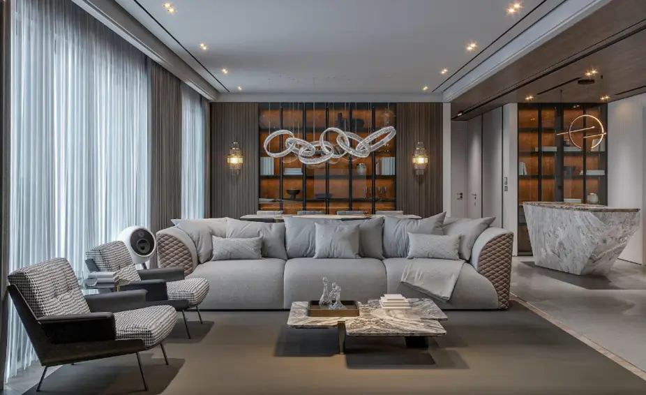 Modern luxury living room with a large grey sofa, accent chairs, marble coffee table, statement lighting, and built-in shelving.