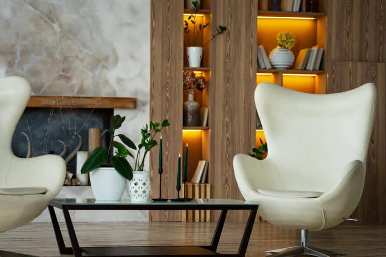 Modern living room with two white armchairs, a glass coffee table, plants, and illuminated shelves with books and decor.