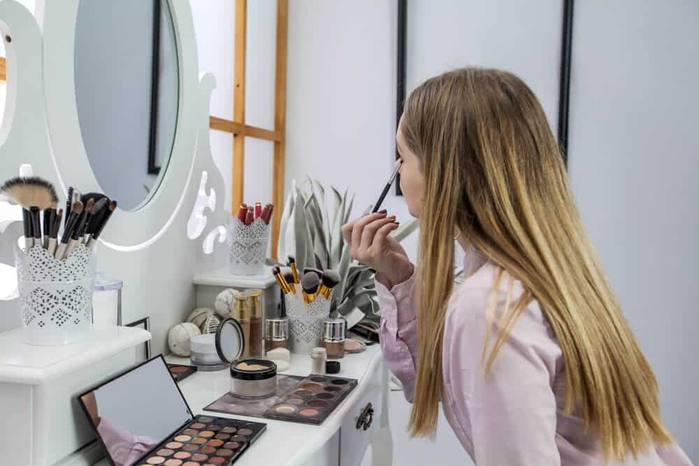 A woman applies eyeliner while looking in a mirror on a vanity table filled with various makeup products, preparing for the day after a restful night surrounded by her favorite mattresses & sleep essentials.