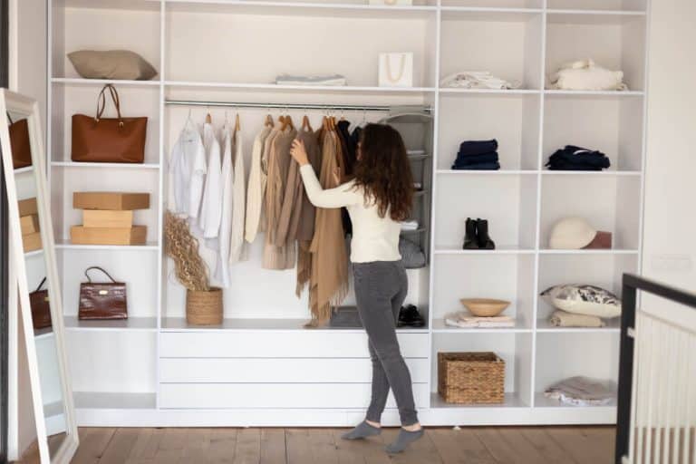 A woman with long brown hair stands in front of an open closet, organizing clothes on hangers. The closet has neatly arranged shelves filled with folded clothes, bags, boxes, and shoes. Nearby, a selection of mattresses & sleep essentials adds to the serene vibe. She is dressed in a light sweater and gray jeans.