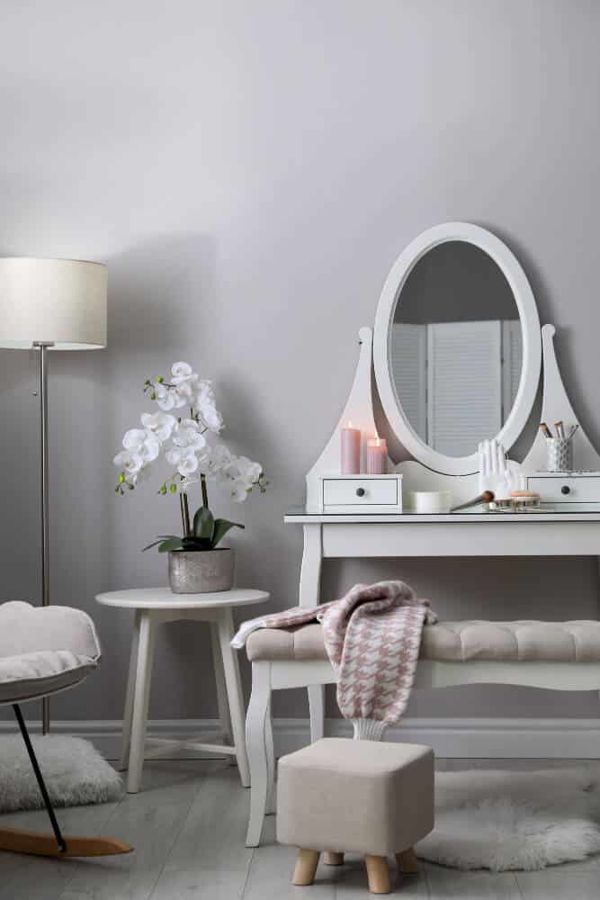 A white vanity table with a mirror, candles, a potted orchid, a cushioned bench, and a small stool in a cozy room filled with mattresses and sleep essentials.