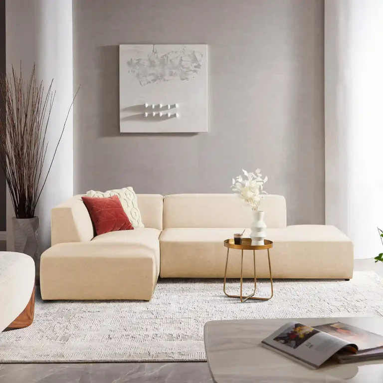 L-shaped beige sofa with a red accent pillow, gold side table, and decorative vase in a modern living room setting.