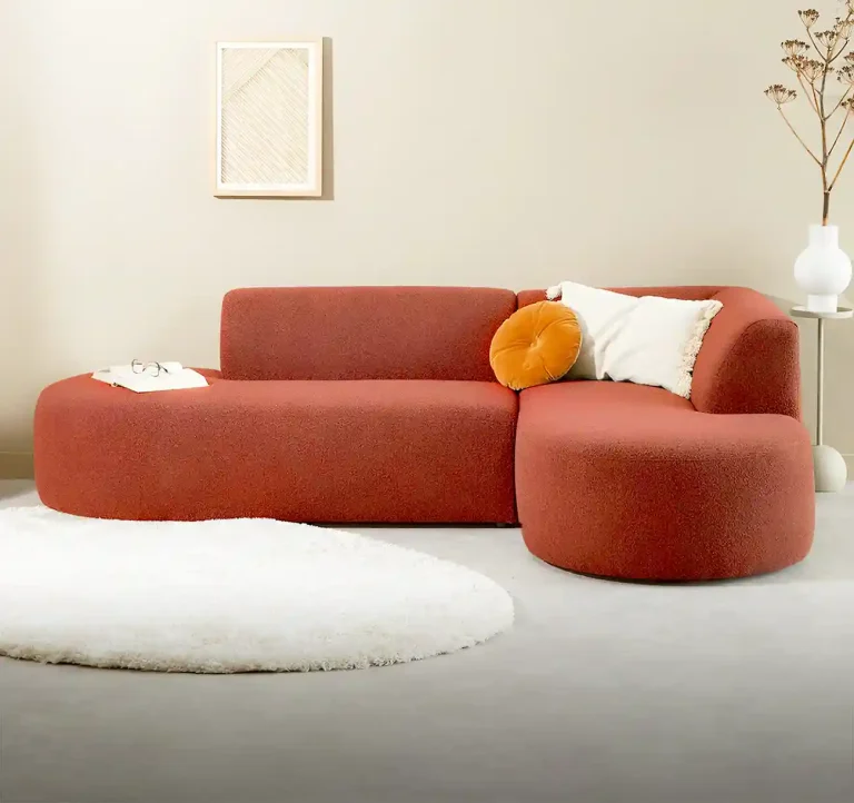 Rust-colored sectional sofa with rounded edges, a decorative pillow, and a soft white rug in a modern living space.