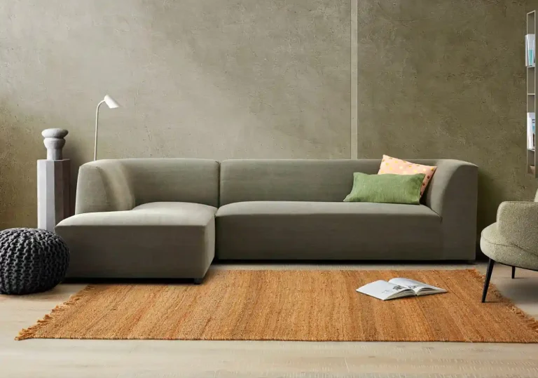 Cozy living room featuring a green sectional sofa, colorful cushions, a knitted pouf, and a wooden floor rug with magazines.