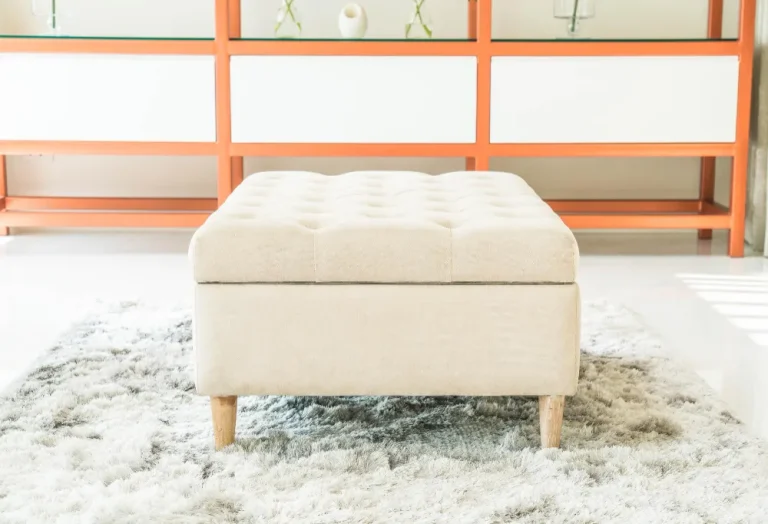 A white ottoman placed on a rug, positioned in front of a vibrant orange wall.