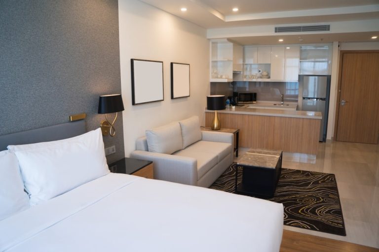 A modern hotel room with a bed, sofa, and kitchenette. The room features a neutral color scheme with wooden accents and includes two framed artworks and contemporary lighting.