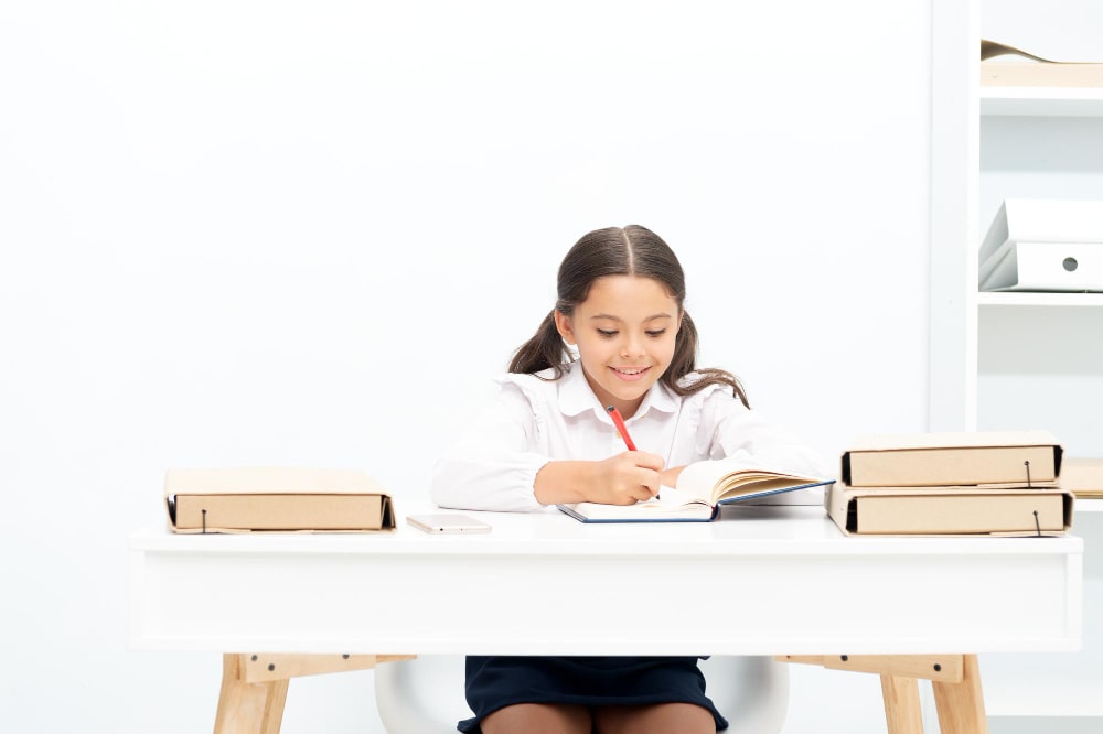 A girl sits at a desk, focused on a book, with a pen in hand, engaged in her studies.