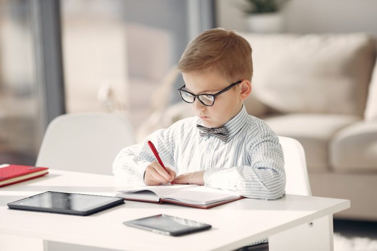 A young boy wearing glasses is focused on writing in his notebook, showcasing his concentration and creativity.